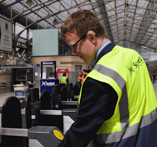 A young man in a high-vis jacket operates a ticket barrier in a train station