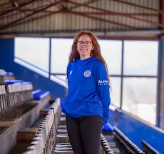 A young woman stands next to a row of seats in a stand within Palmerston Park.