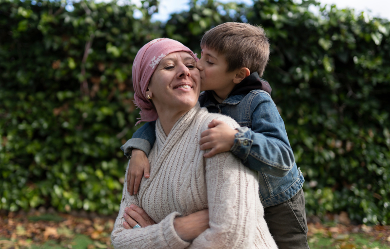 Young boy in denim jacket kissing his smiling mother wearing a pink headscarf outdoors with green foliage background.