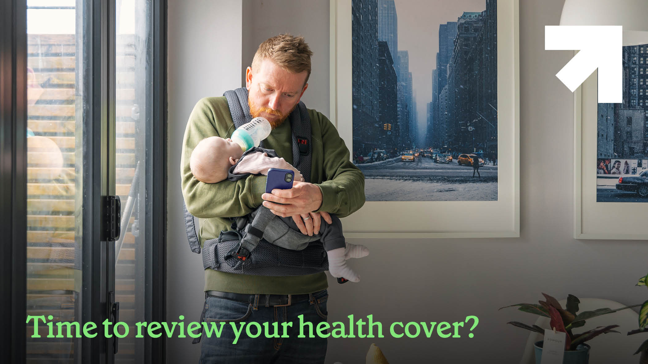 Man holding and feeding is baby, while scrolling on his phone. Text reads "Time to review your health cover?"