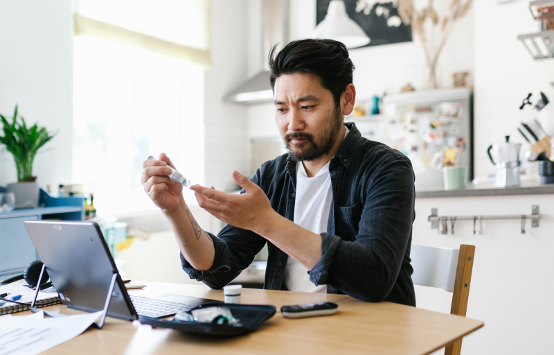 Man with dark hair and beard checking blood glucose level with testing device at home desk with laptop in bright kitchen