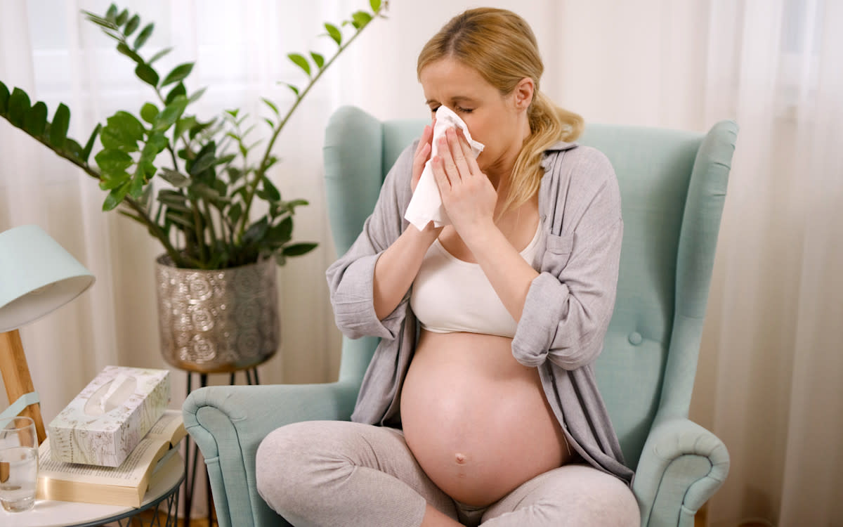 A pregnant woman sitting on a lounge blowing her nose.