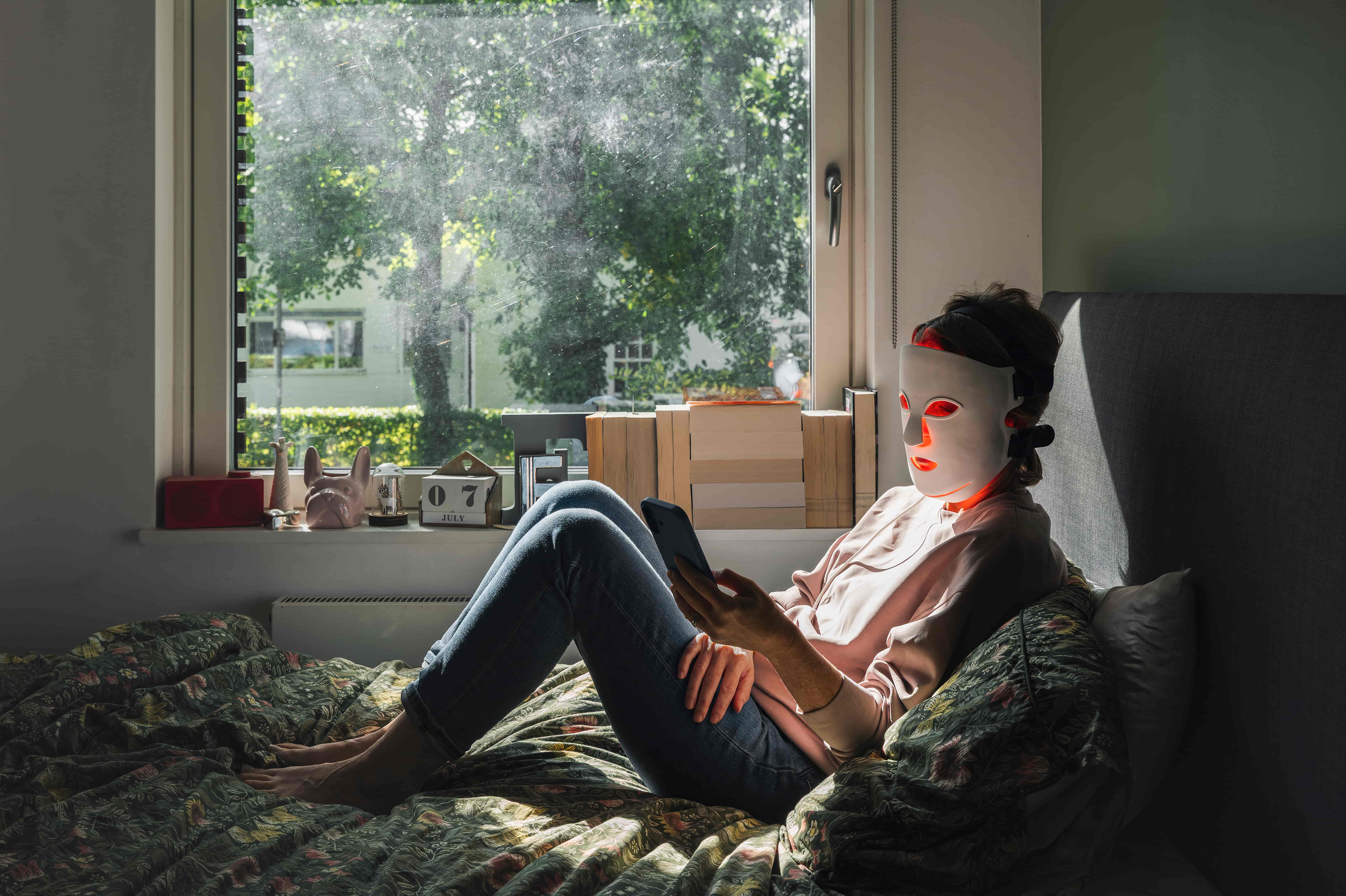 A woman wears a red-light therapy mask while sitting on her bed in a well lit room