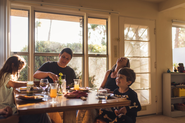 A well lit dining room has a full table of cups and plates, while two children sit eating dinner with their mum and dad