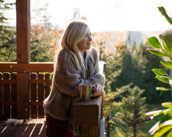 Woman in beige cardigan holding mug on sunny deck overlooking trees and hills in natural daylight