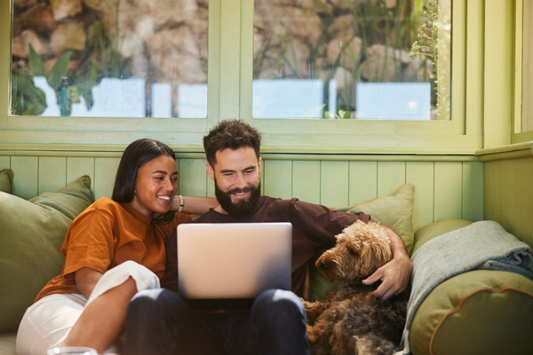 Young couple and fluffy dog sitting on a lounge looking at a laptop screen