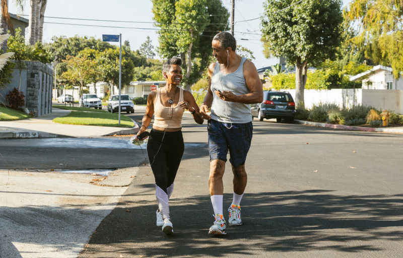 Two people jogging together on a sunny suburban street lined with trees and parked cars.