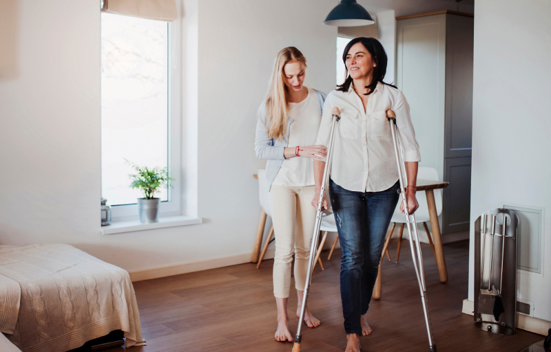 Woman on crutches being assisted by carer in bright modern home with wooden floors and neutral tones