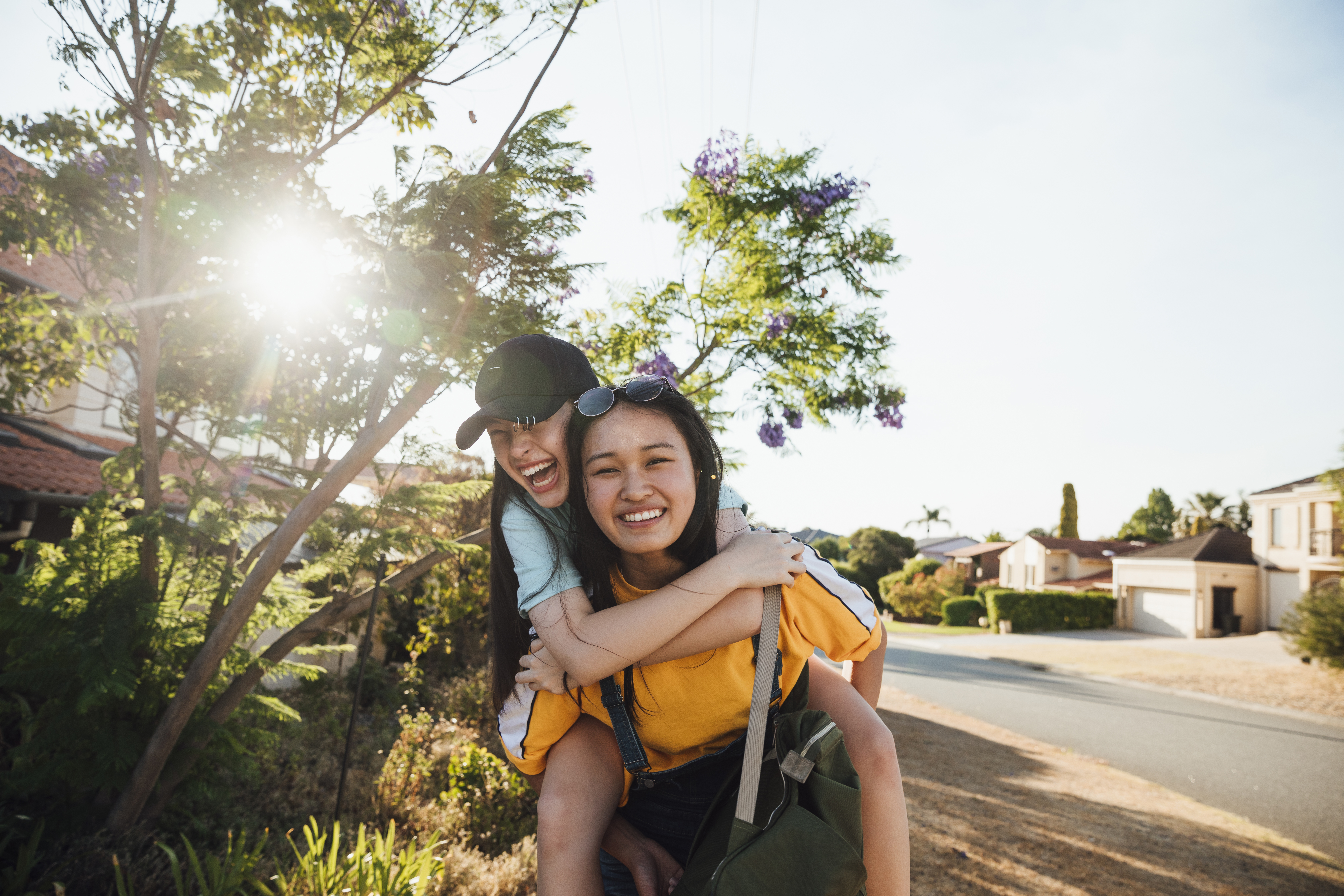 Teenage girls hanging out together walking along a neighbourhood street. One girl is giving the other a piggyback as they laugh together enjoying summer.