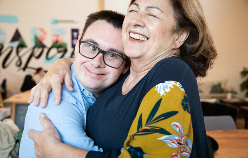 A smiling woman in a floral sleeve top embraces a young man wearing glasses and a blue shirt in a warm hug indoors.