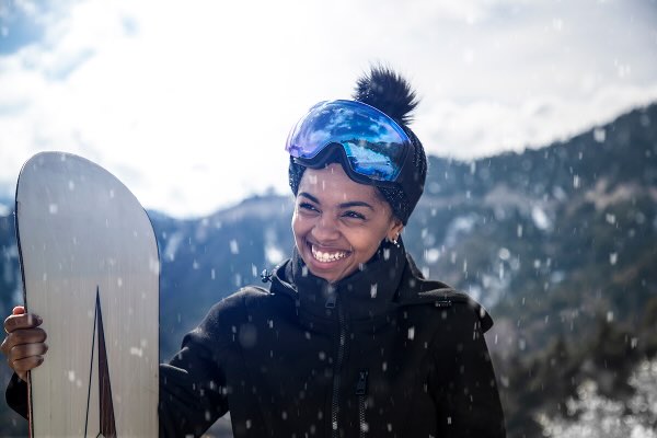 A smiling woman in the snow wearing snow googles and a beanie, holding her snowboard