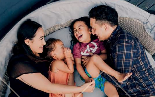 Two adults and two children lying together in a circular outdoor lounge or tent, facing one another in a relaxed and playful moment. One child is wearing a pink top and the other an orange top, while the adults are positioned on either side. The group is closely gathered, creating a warm family scene.