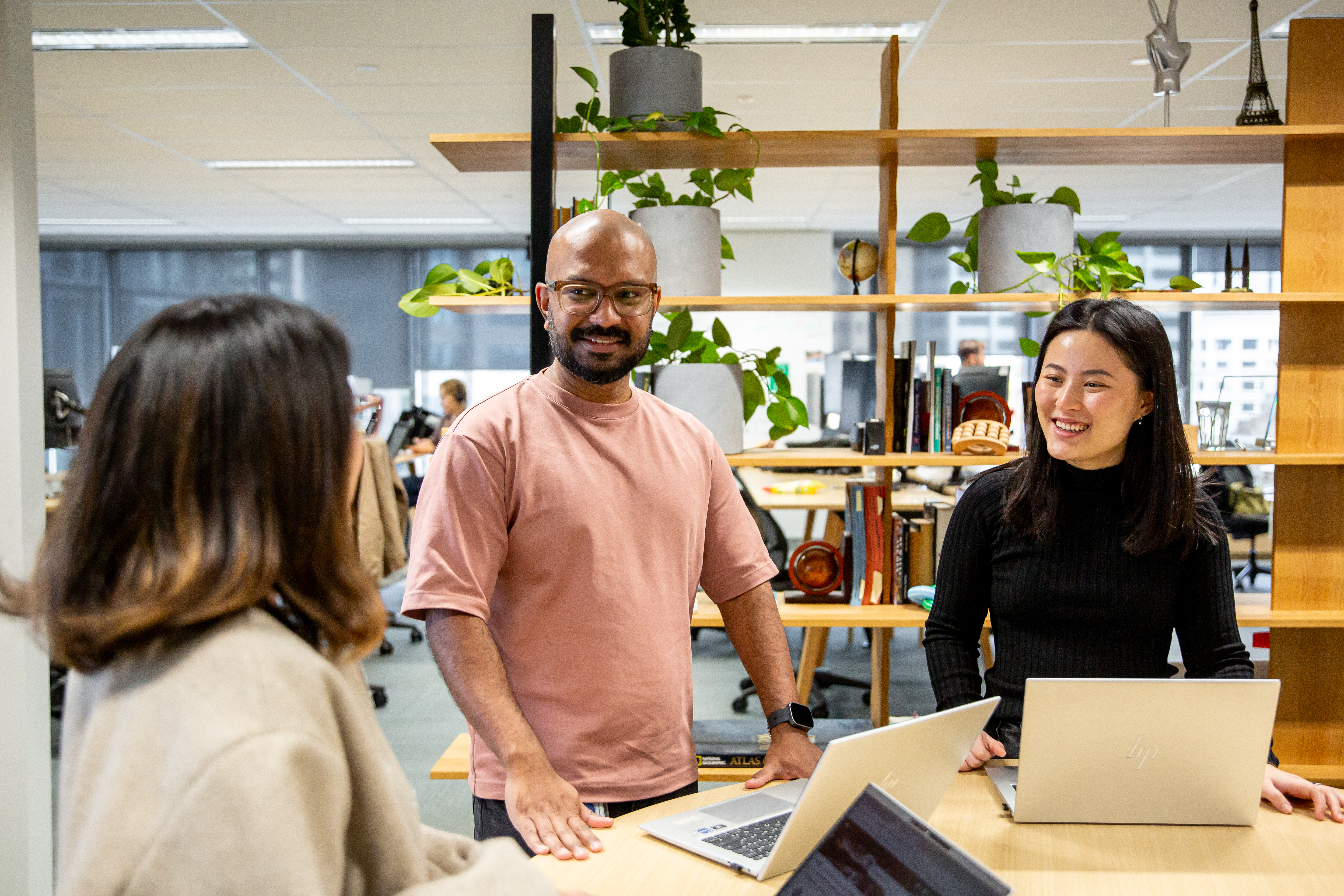 Three people collaborating in a modern office workspace. Two people are standing and seated behind open laptops, while a third person is visible in the foreground with their back to the camera.