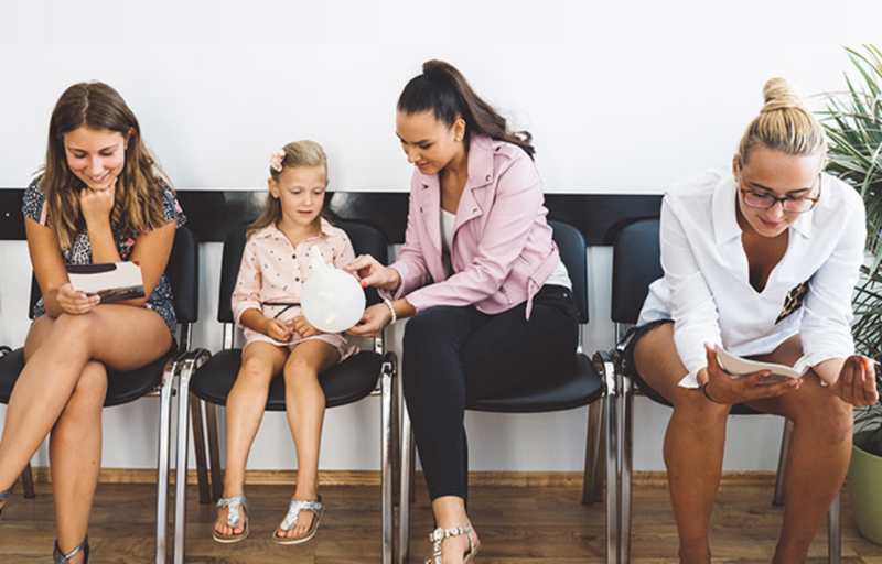 Four people sitting in a waiting room: two women looking at medical papers, a child holding an inflated glove, and a woman in pink assisting.
