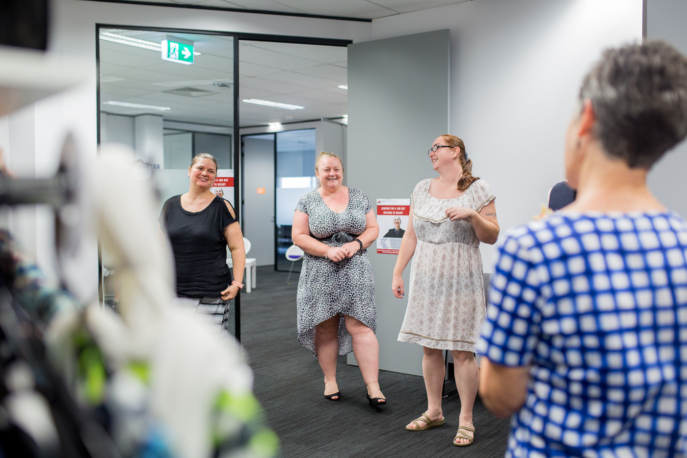Four women standing and smiling in a modern office space with glass doors and grey walls during a workplace gathering.