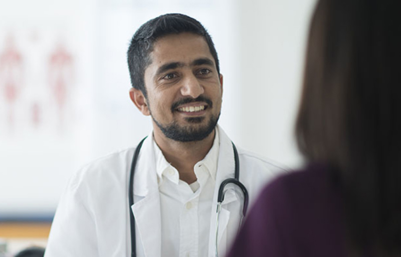 Smiling male doctor in white coat with stethoscope speaking with patient in medical consultation room