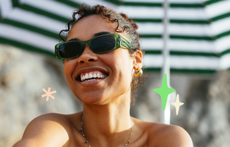 Woman smiling with sunglasses on at the beach under an umbrella.