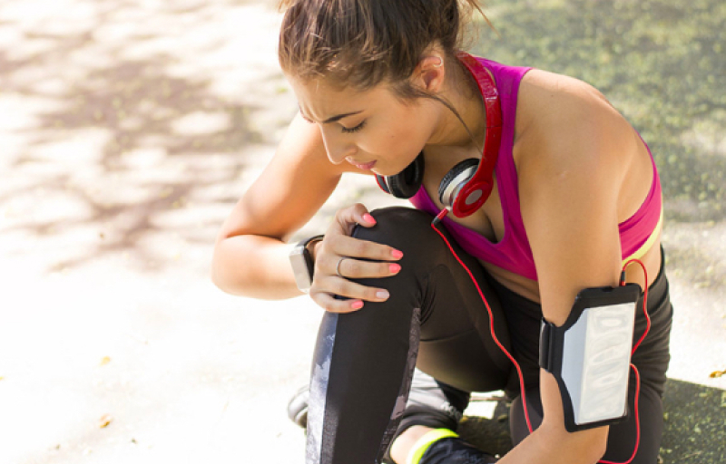 Woman wearing pink sports bra and headphones around her neck, holding injured knee on the ground