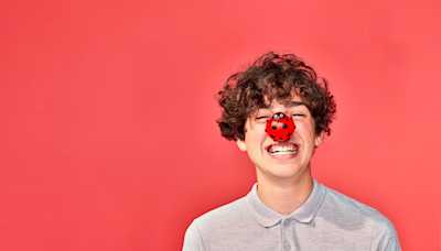 A boy laughing and wearing a red nose in the shape of a ladybird.