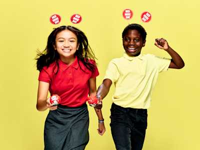 Two young people racing in a nose and spoon race and wearing Red Nose Day antennae