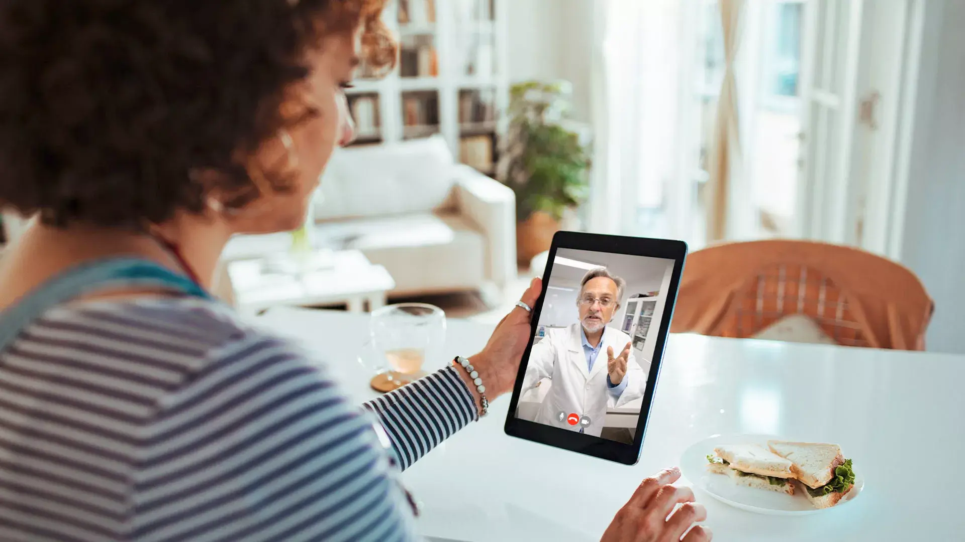 Vrouw zit aan tafel in woonkamer en facetimet dokter op tablet