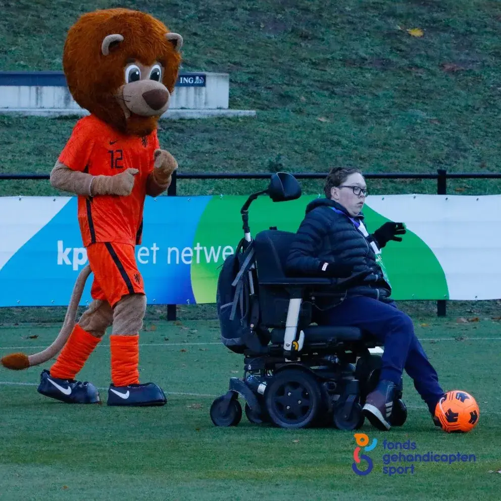 Jongen in rolstoel trapt ee nvoetbal terwijl de KNVB leeuw erachter staat aan te moedigen.