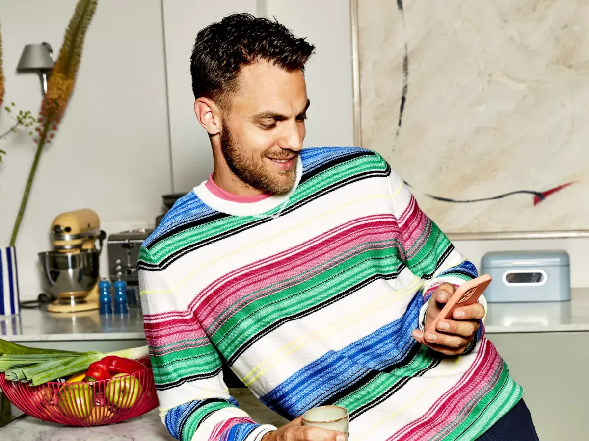 Jongeman met gekleurde trui leunt tegen keukenblad met koffie en mobiel in zijn handen
