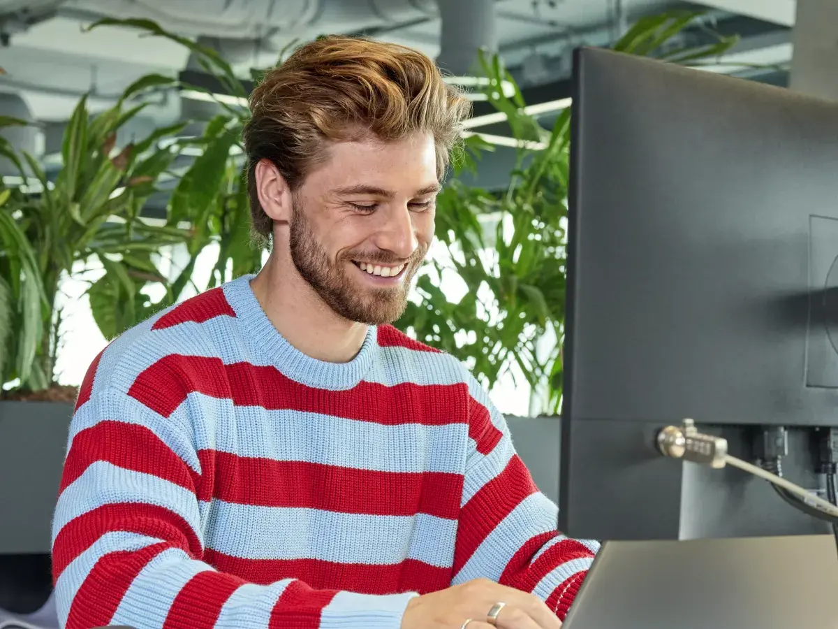 Een man werkt aan zijn bureau op zijn laptop en computerscherm. Hij kijkt lachend naar zijn laptop.