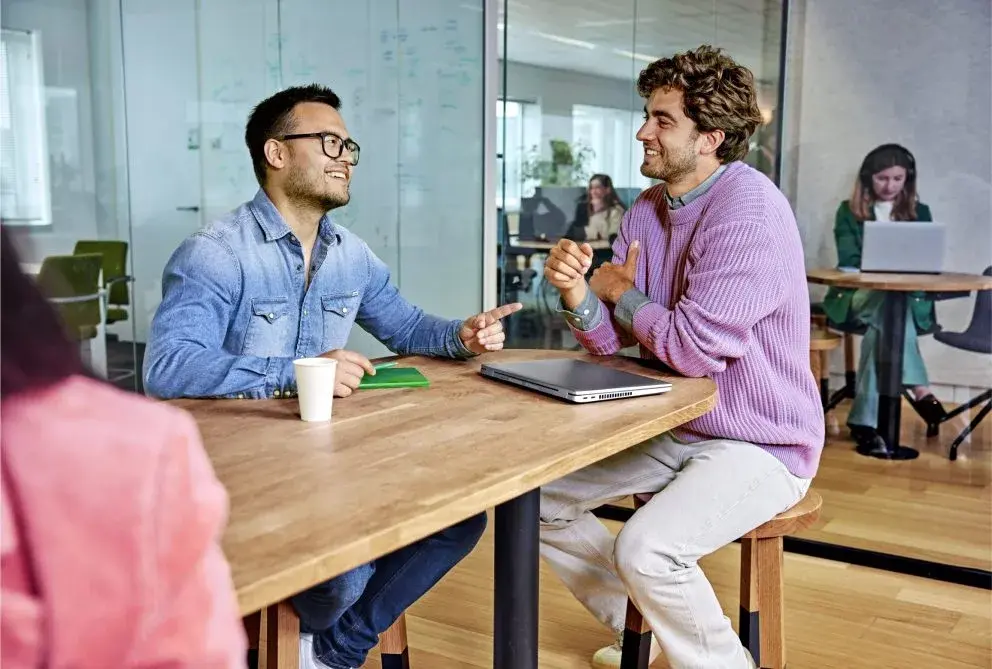 twee mannen zitten aan een tafel in overleg in een ruimte waar meerdere mensen werken