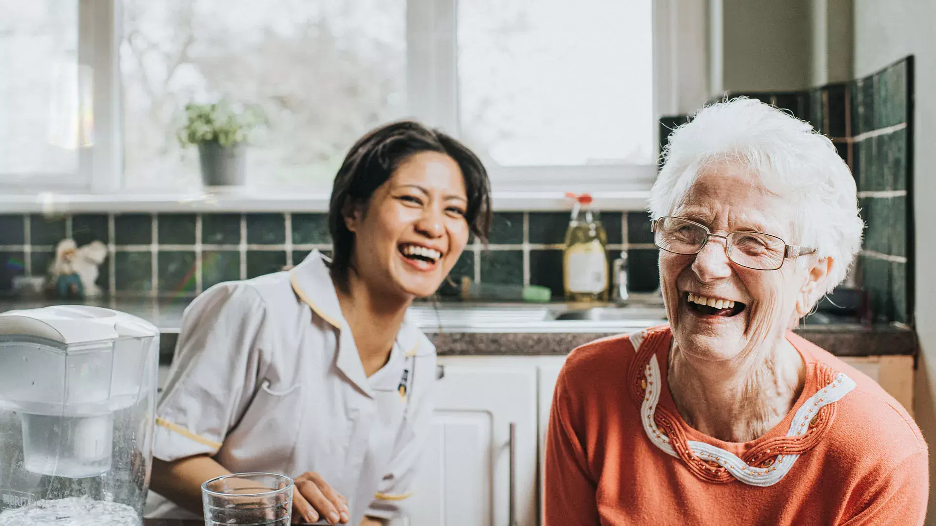Zorgverlener en oudere vrouw zitten in de keuken en lachen met elkaar