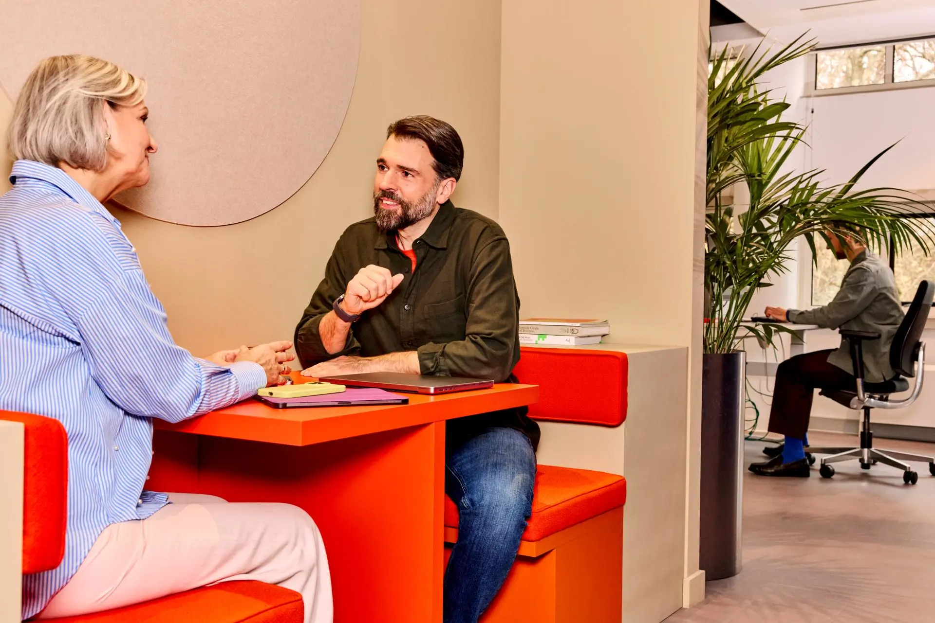 Twee collega's zitten samen en man aan bureau op de achtergrond