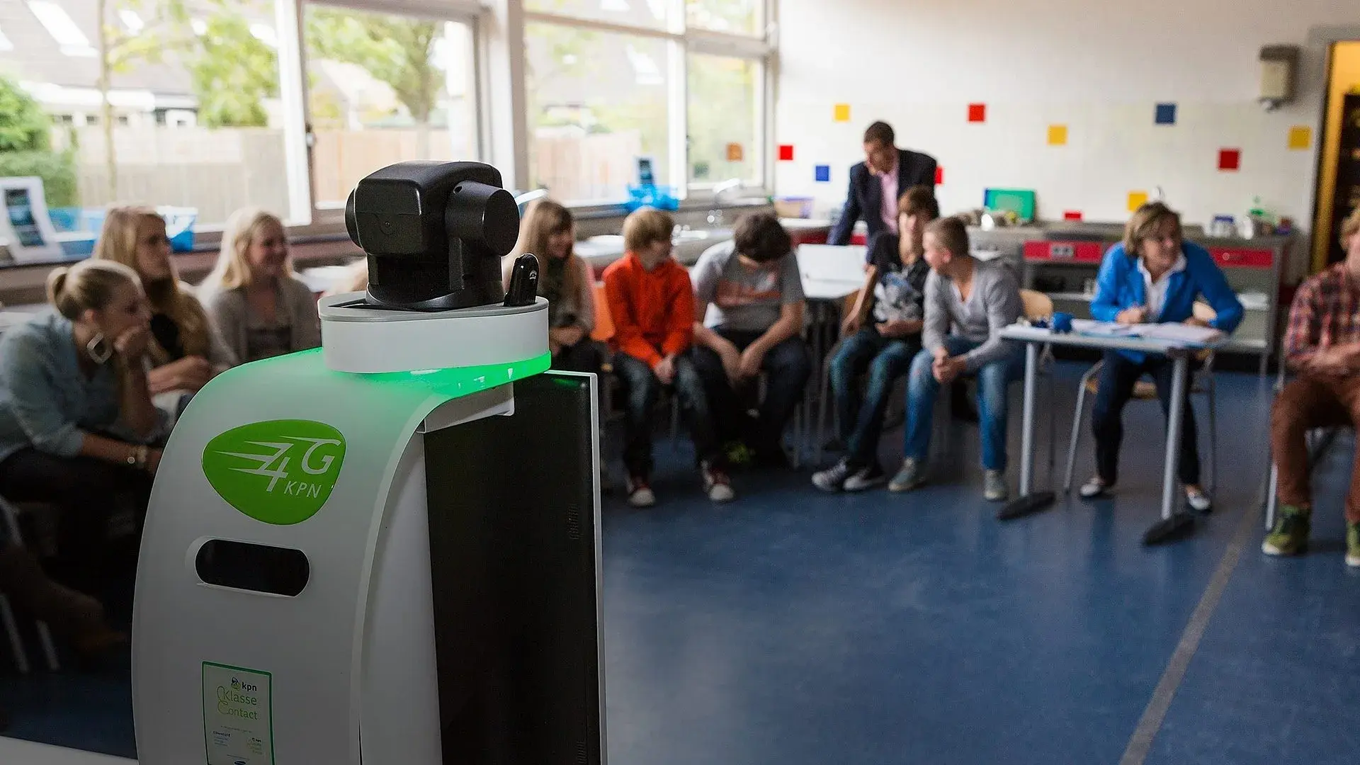 Kinderen in de klas in kringgesprek met de nieuwe klasgenoot op voorgrond waarop het zieke kind thuis mee kan doen met kringgesprek