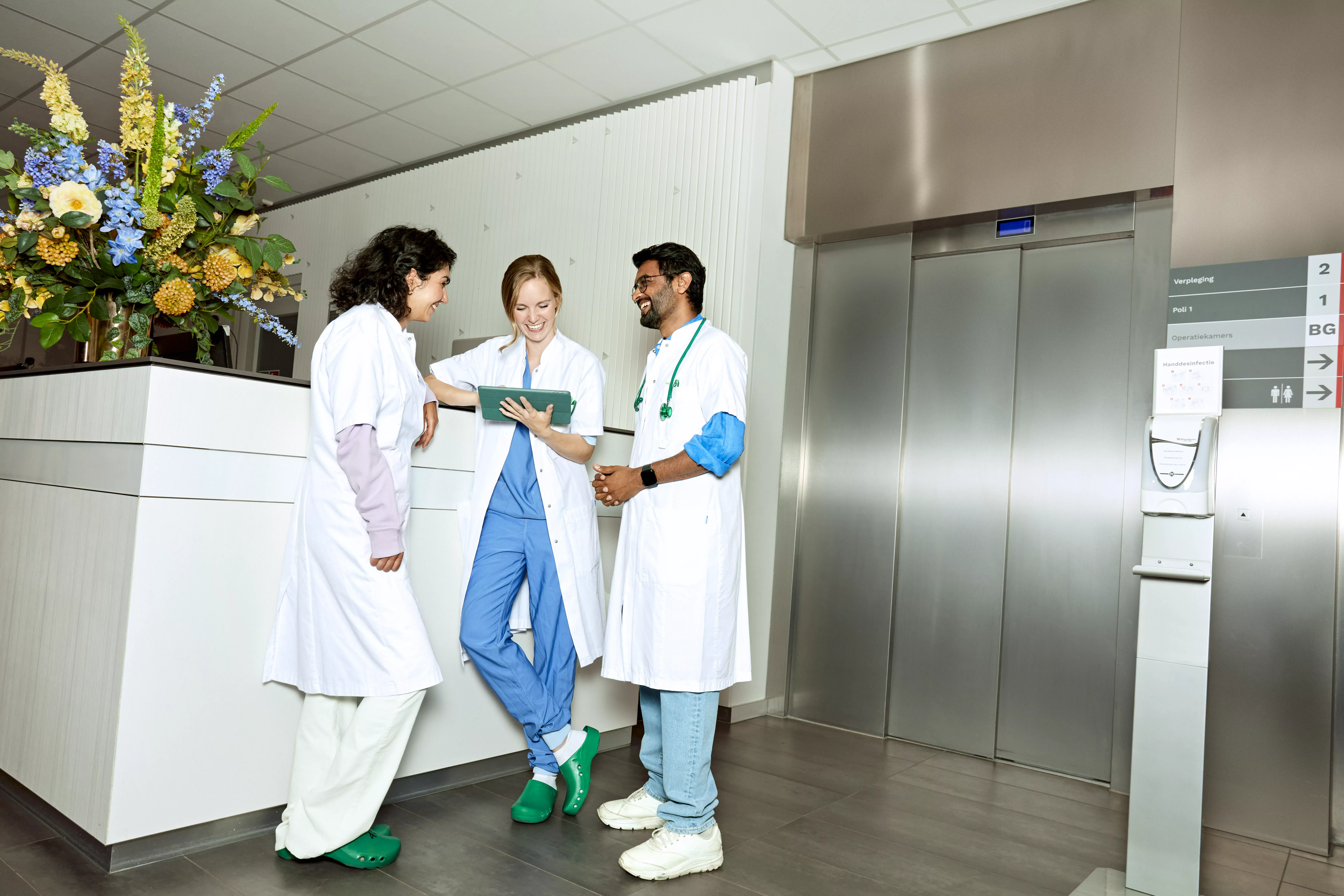 Drie artsen staan lachend aan de balie van een ziekenhuis met een tablet