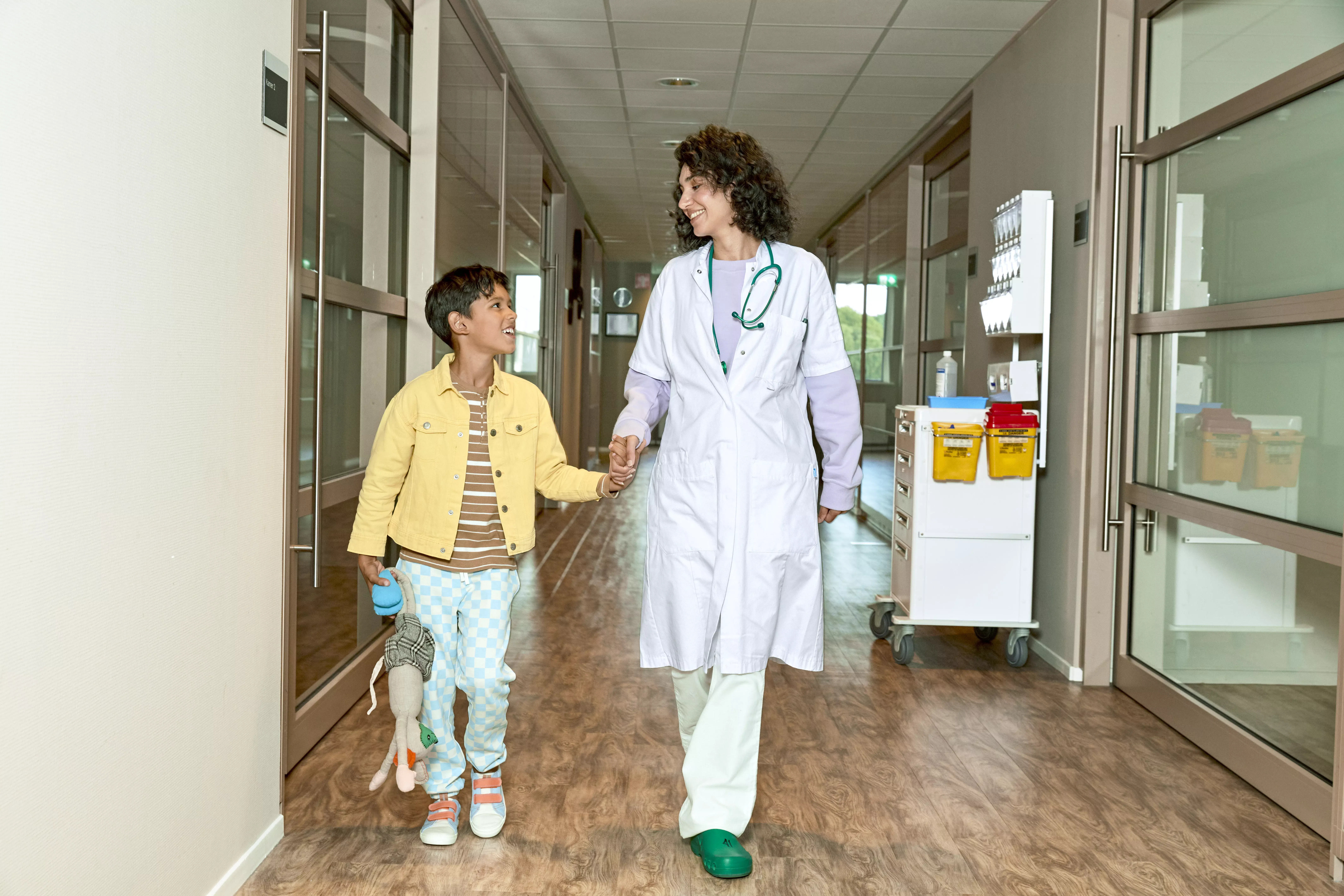 Jongetje en vrouwelijke arts lopen hand in hand door een ziekenhuisgang