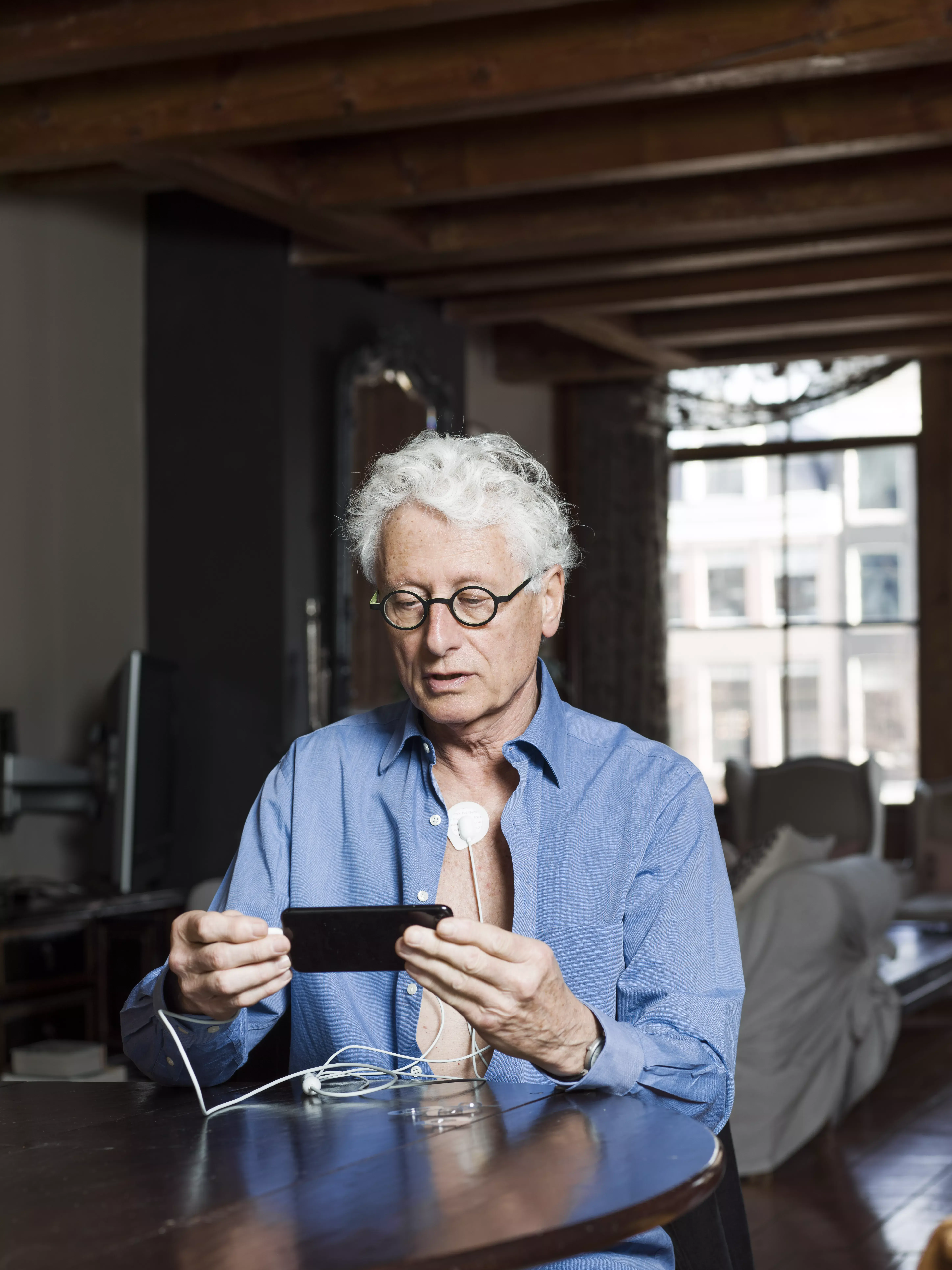 Oudere man met bril zit aan tafel en kijkt op zijn telefoon
