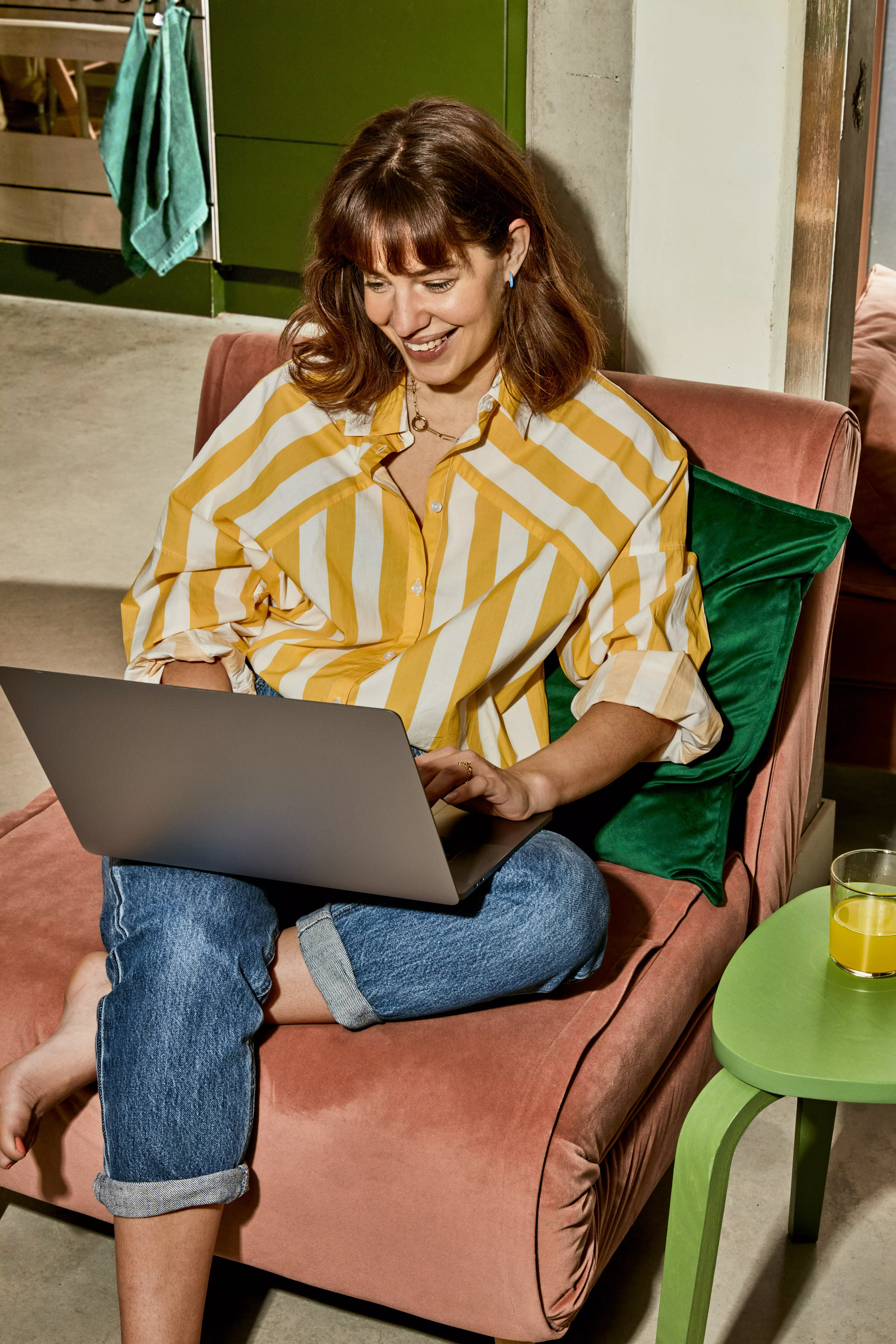 Een vrouw zit op een bank met haar laptop op schoot. Ze lacht en kijkt naar haar laptop.