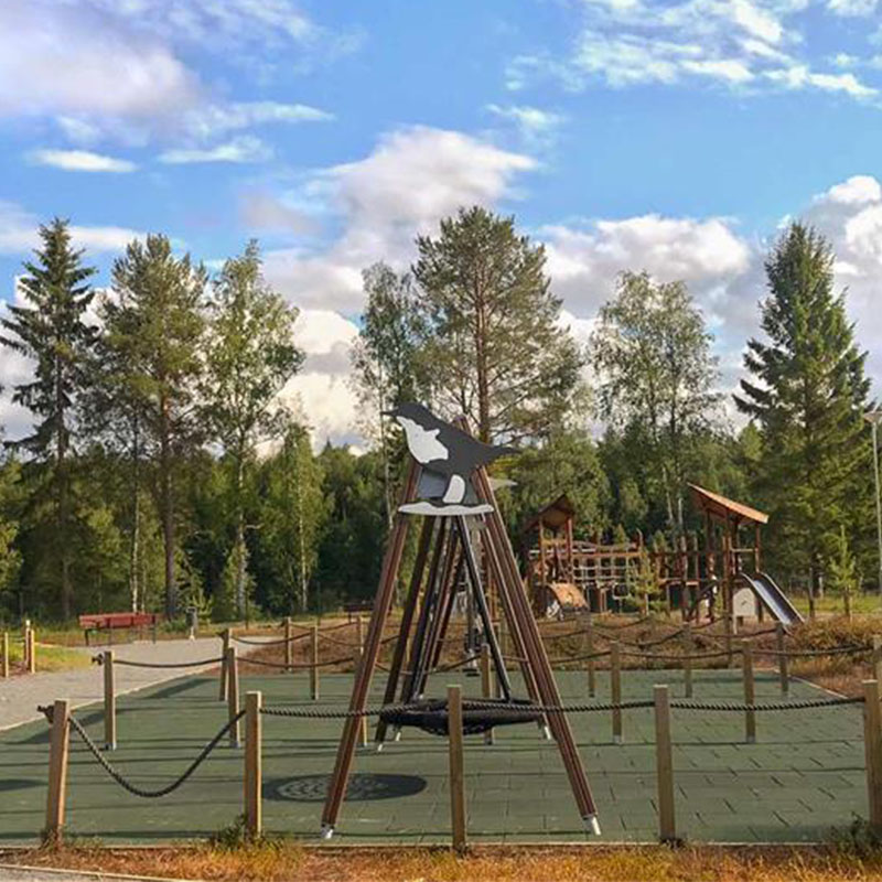 Flora Nature Play Ii Koskikara, Finland A children's playground surrounded by trees under a partly cloudy sky. The playground features various structures, including a large swing set with ropes and a decorative bird figure on top, as well as climbing equipment in the background.