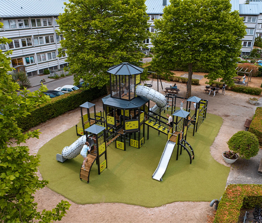 Outdoor Playground in Denmark: Text Liftup 2 A children's playground with multiple climbing structures, a slide, and several interconnecting tunnels is set on green artificial turf. The play area is surrounded by trees and adjacent to residential buildings with people sitting at picnic tables in the background.