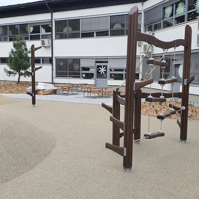 Playground of Škofljica Primary School A school playground features a climbing structure with ropes and wooden steps on a beige rubber surface. In the background, there are benches and a modern building with large windows adorned with floral decorations. A small tree is visible on the left side.