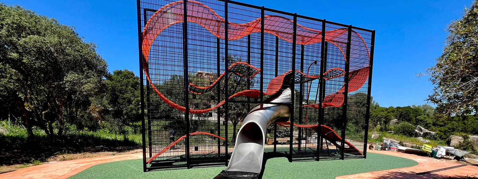 Wall-Holla Park Sardinia Image Liftup A unique playground structure features a tall metal framework with an intricate red mesh design. A silver tunnel slide extends from the framework to the green ground below. Trees and a clear blue sky serve as the backdrop.