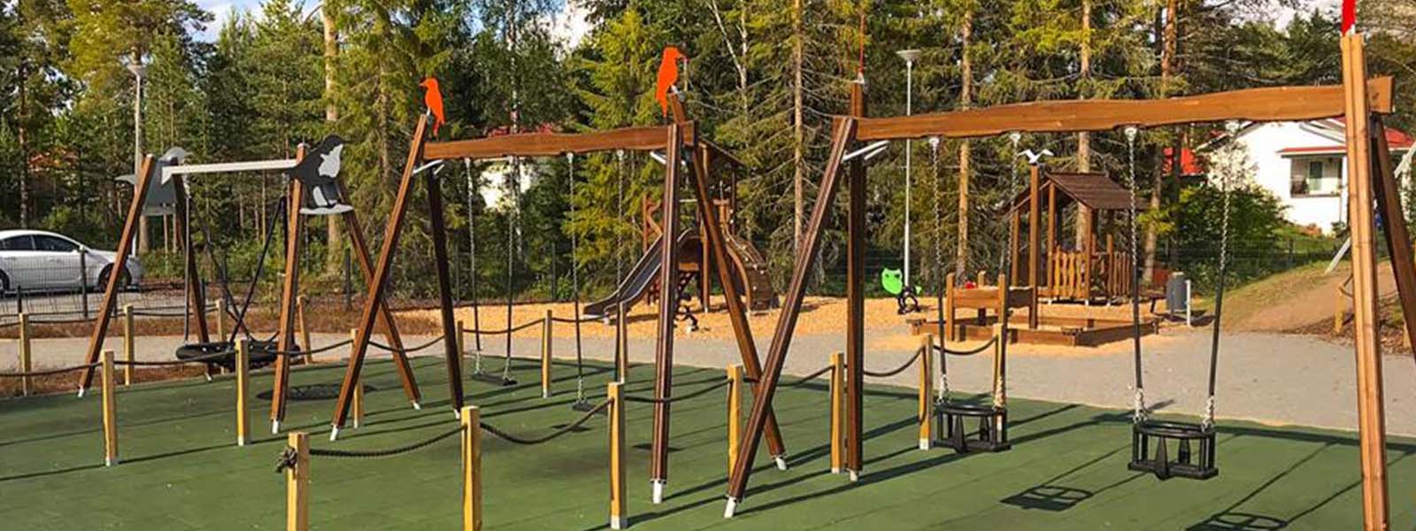 Flora Nature Play Ii Koskikara, Finland A spacious playground with various activities, including swings, balance beams, and climbing structures. The playground features green flooring, surrounded by trees and wooden picnic benches. There is a parked car to the left, and houses are visible in the background.