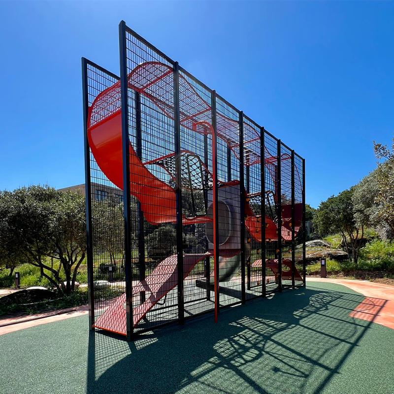 Wall-Holla Park Sardinia 1 A playground structure with a red spiral slide enclosed in black mesh netting. The structure stands on a rubberized play surface under a clear blue sky. Trees and grassy areas surround the playground.