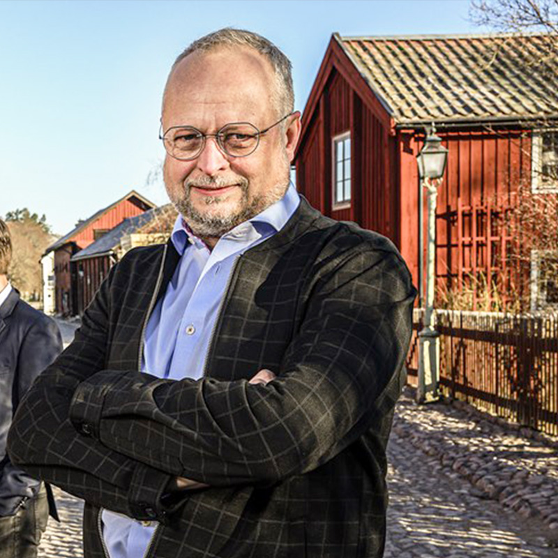 VD Magnus LasseMaja 800x800 A man with glasses and a beard stands with arms crossed, smiling confidently. He is wearing a blue shirt and a dark plaid jacket. Behind him, there is a traditional wooden building painted in red, and the scene appears to be in a quaint village.