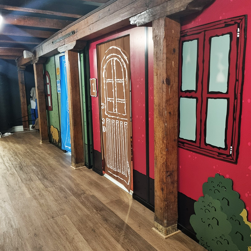 Rio och Aaron LasseMaja 4 800x800 A hallway with a wooden floor and timber beams features colorful, cartoon-style wall art. The illustrations include green and red walls, windows, and doors, one of which is brown with a white, hand-drawn design. Bushes are painted near the bottom.