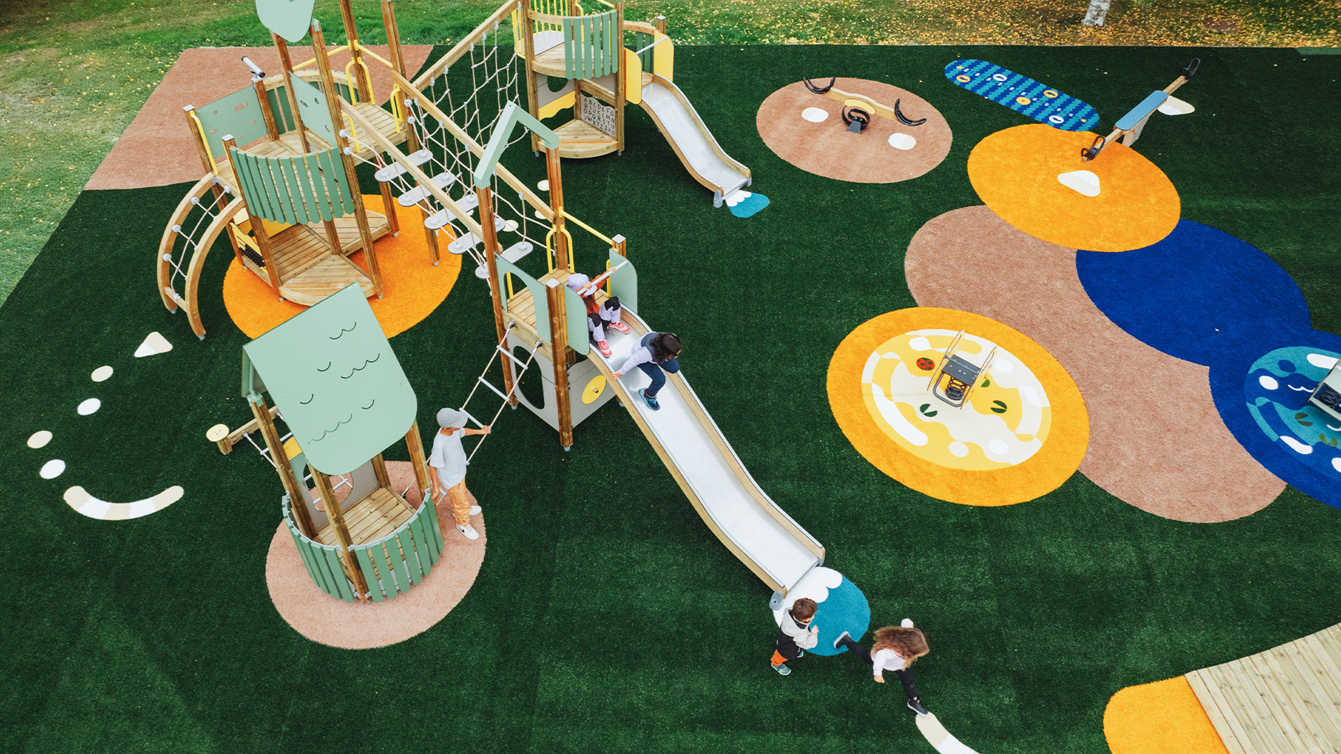Children playing and sliding in a Lappset Prime playground Aerial view of a Lappset Prime playground where children are playing, climbing, and sliding. The area features a colorful safety surface and green artificial grass.