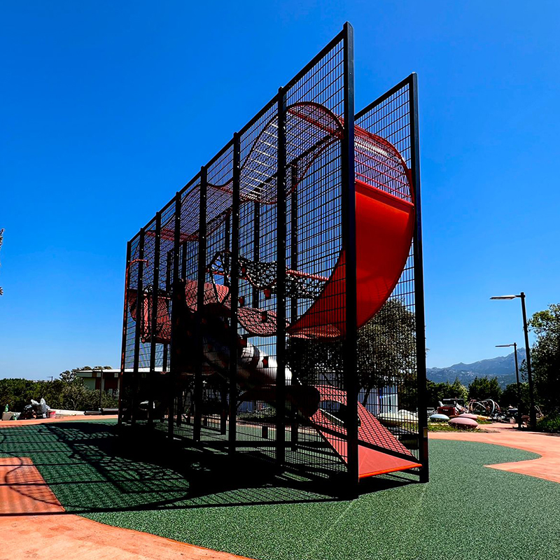 Wall-Holla Park Sardinia 2 A playground structure with a red curvy slide is encased in black mesh fencing. The structure is set on turf with a clear blue sky in the background. There are trees and additional park equipment visible.
