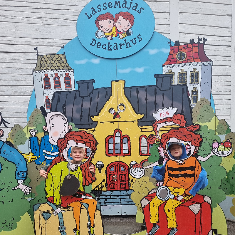 Rio & Aaron on LasseMaja's detective agency Children smile through face cut-outs in a colorful board at LasseMajas Deckarhus, a detective-themed attraction. The board features cartoon characters in a whimsical town setting with magnifying glasses, a yellow house, and playful details.