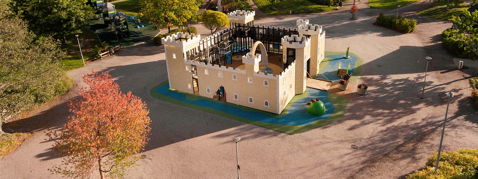 Familypark in Lahti Finland Aerial view of a playground featuring a large, castle-shaped play structure with yellow walls and turrets. The surrounding area includes green trees, a few park benches, and colorful ground surfaces. Pathways wind around the playground under a clear sky.