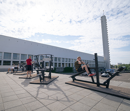 SIsu Fitness Sports area in Helsinki Olympic stadium People are seen working out on exercise equipment set up on a spacious outdoor rooftop gym. The setting is bright with daylight, and a tall, white building with a tower is visible in the background against a partly cloudy sky.