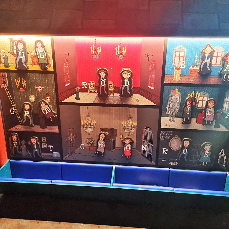 Rio och Aaron LasseMaja 7 800x800 A detailed wall display features various puppet figures in different settings, all related to letters forming the word "GRIMM." The colorful scenes depict characters in medieval attire, surrounded by intricate props and furnishings. Blue seating is visible in front.